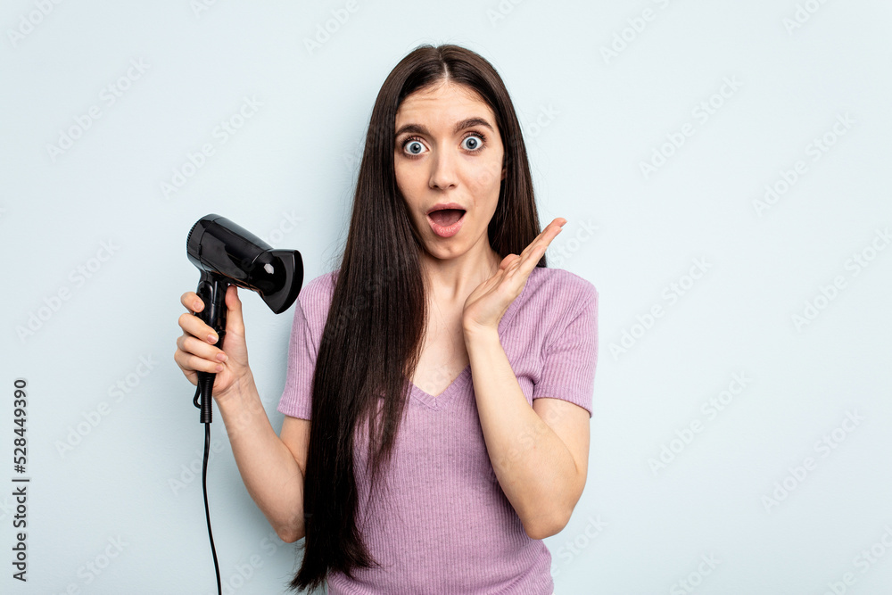 Young caucasian woman holding hairdryer isolated on blue background surprised and shocked. Young caucasian woman holding hairdryer isolated on blue background surprised and shocked.