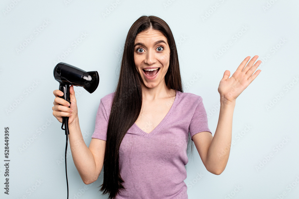 Young caucasian woman holding hairdryer isolated on blue background receiving a pleasant surprise, excited and raising hands. Young caucasian woman holding hairdryer isolated on blue background receiving a pleasant surprise, excited and raising hands.