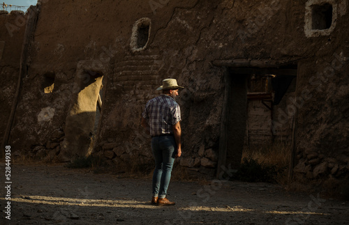 Rear view of adult man in cowboy hat and shirt against abandoned building