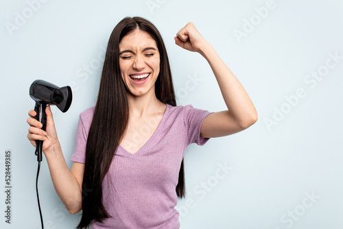 Young caucasian woman holding hairdryer isolated on blue background raising fist after a victory, winner concept.