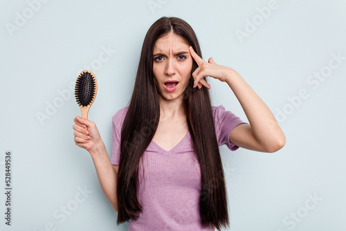 Young caucasian woman holding a hairbrush isolated on blue background showing a disappointment gesture with forefinger.