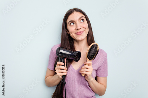 Young caucasian woman comb your hair with the dryer isolated on blue background