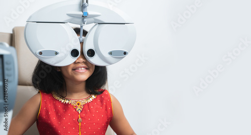 Happy Indian child girl doing subjective refraction with phoropter digital modern machine for eyesight test at hospital or optometry clinic. Copy space. White background photo