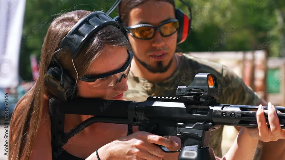 Caucasian woman in safety headphones and goggles aiming submachine gun watched by bearded man in camouflage t-shirt. Firearms training ar fiiring range. Horizontal shot. High quality 4k footage Caucasian woman in safety headphones and goggles aiming submachine gun watched by bearded man in camouflage t-shirt. Firearms training ar fiiring range. Horizontal shot. High quality 4k footage