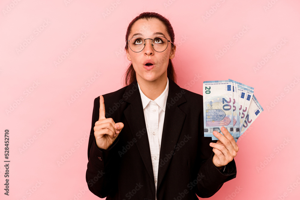 Young business caucasian woman holding bank notes isolated on pink background pointing upside with opened mouth. Young business caucasian woman holding bank notes isolated on pink background pointing upside with opened mouth.