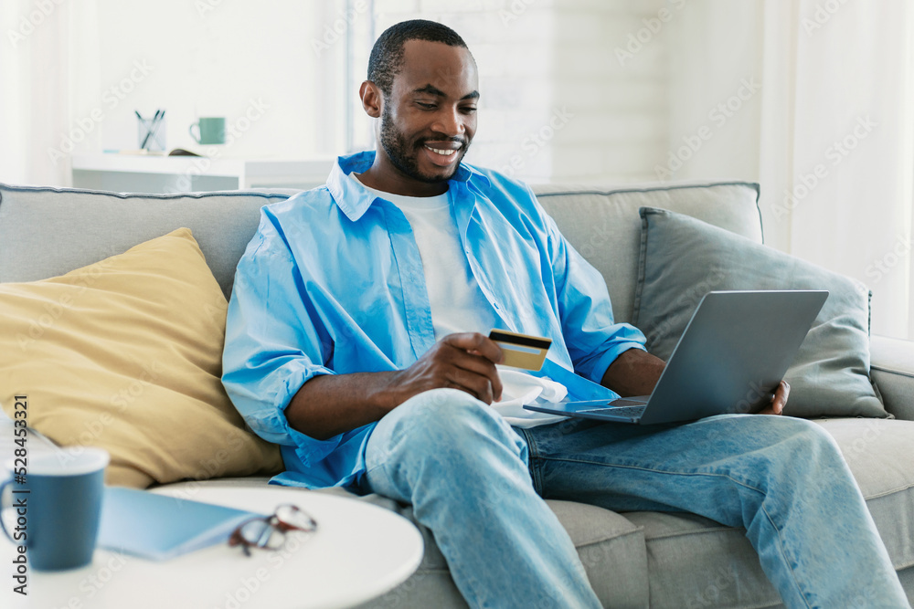 Happy african american man holding credit card in hand and using laptop computer, making financial transaction at home Happy african american man holding credit card in hand and using laptop computer, making financial transaction at home
