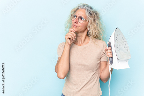 Middle age caucasian woman holding an iron isolated on blue background looking sideways with doubtful and skeptical expression.