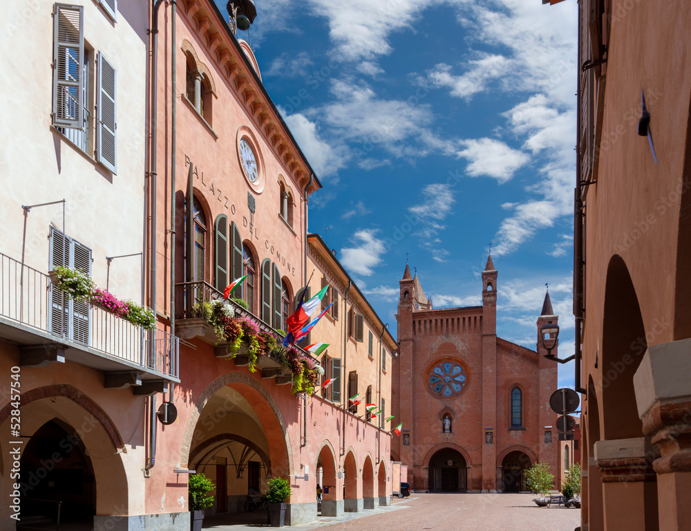 Alba, Langhe, Piedmont, Italy - August 16, 2022: the Town Hall with flowered balconies and medieval arcades in via Cavour, in the background Cathedral of San Lorenzo in Piazza Duomo
