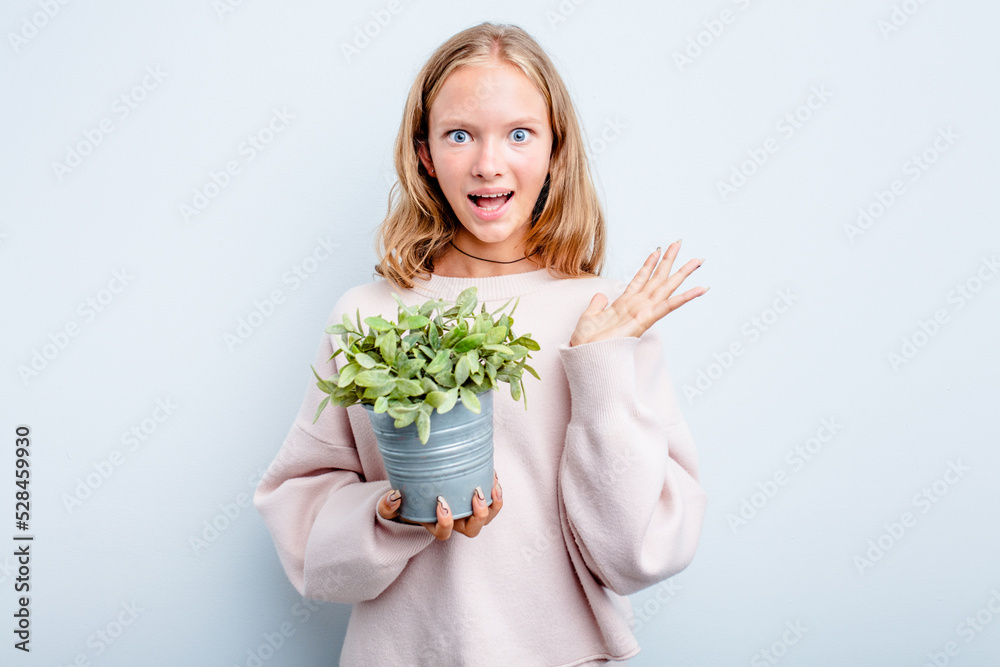 Caucasian teen girl holding a plant isolated on blue background surprised and shocked. Caucasian teen girl holding a plant isolated on blue background surprised and shocked.