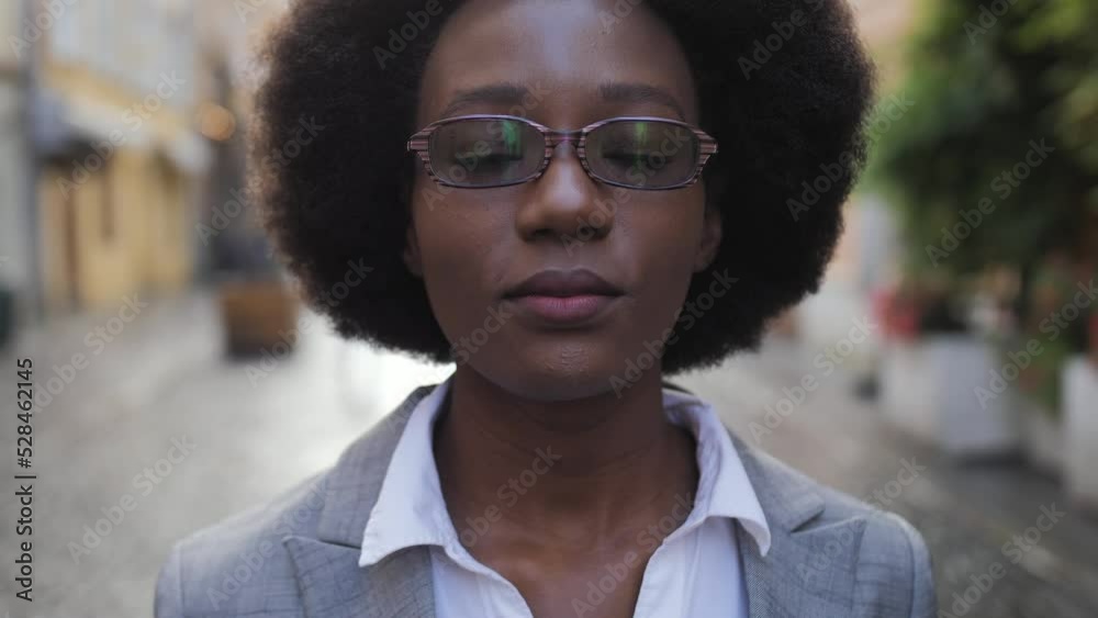 Portrait of positive afro american lady in stylish business suit touching with hand her eyeglasses while standing outdoors. Successful woman smiling and looking at camera on street. Portrait of positive afro american lady in stylish business suit touching with hand her eyeglasses while standing outdoors. Successful woman smiling and looking at camera on street.