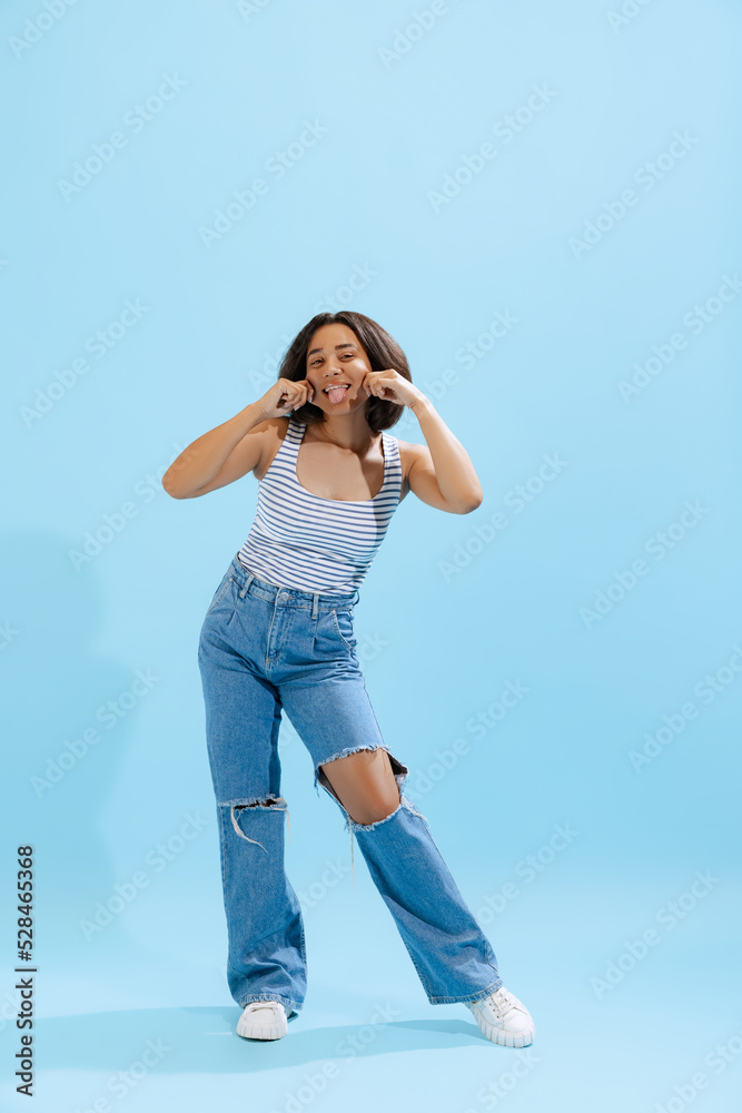 Funny young girl, student wearing casual style clothes having fun, grimaces isolated on blue background. Concept of beauty, art, fashion, human emotions Funny young girl, student wearing casual style clothes having fun, grimaces isolated on blue background. Concept of beauty, art, fashion, human emotions