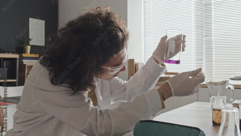 Low angle side-view of curly Biracial guy in white gown, goggles and glove sitting at desk in laboratory, mixing liquid chemicals with dropper in beaker at daytime Low angle side-view of curly Biracial guy in white gown, goggles and glove sitting at desk in laboratory, mixing liquid chemicals with dropper in beaker at daytime