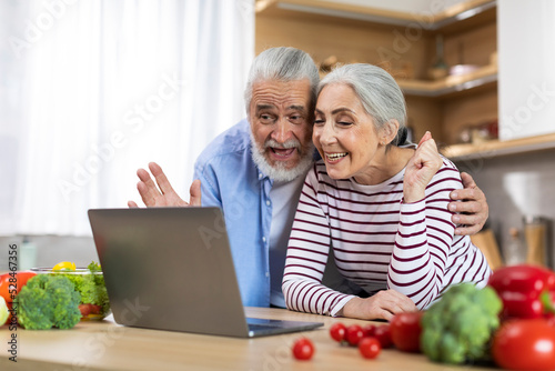 Happy Senior Couple Making Video Call With Laptop While Relaxing In Kitchen