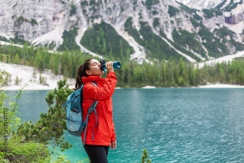 Hydration while on track. Attractive woman hiker in red raincoat drinking water near a lake and mountains