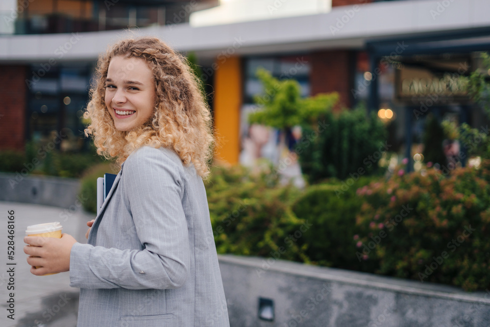 Happy young caucasian woman with curly hair walking down the street turning her head over her shoulder carrying laptop computer, drinking takeaway coffee. Free Happy young caucasian woman with curly hair walking down the street turning her head over her shoulder carrying laptop computer, drinking takeaway coffee. Free