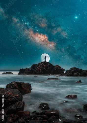 silhouette of person on the top of the rock with moon and milky way in the background over the ocean