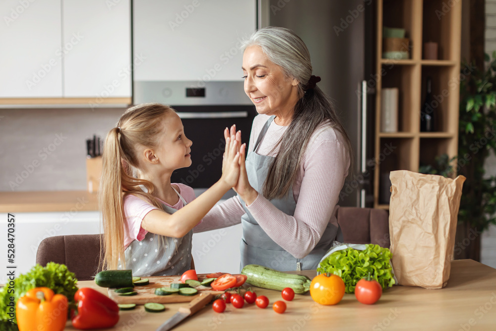 Smiling european small girl and old grandmother preparing salad from organic vegetables, touching with hands Smiling european small girl and old grandmother preparing salad from organic vegetables, touching with hands