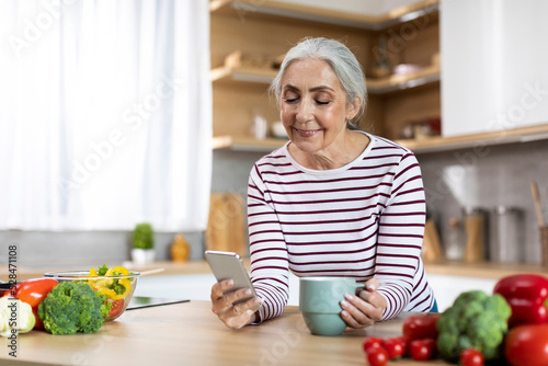 Portrait Of Smiling Senior Woman Using Smartphone And Drinking Coffee In Kitchen
