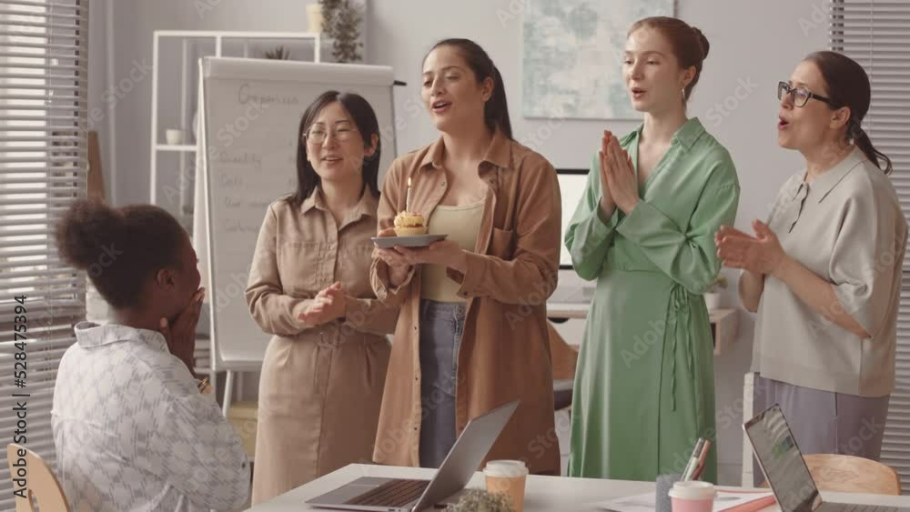 Medium slowmo of group of young cheerful women with Birthday cake singing Happy Birthday to their African American female colleague
