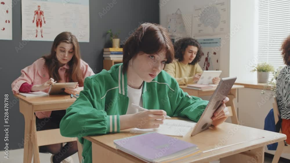 Chest-up of short-haired Caucasian girl sitting at desk on anatomy lesson, using tablet computer in afternoon Chest-up of short-haired Caucasian girl sitting at desk on anatomy lesson, using tablet computer in afternoon
