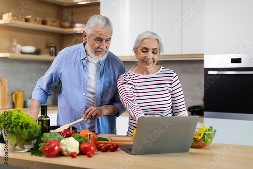 Cheerful Elderly Spouses Looking Recipe On Laptop While Cooking Food In Kitchen