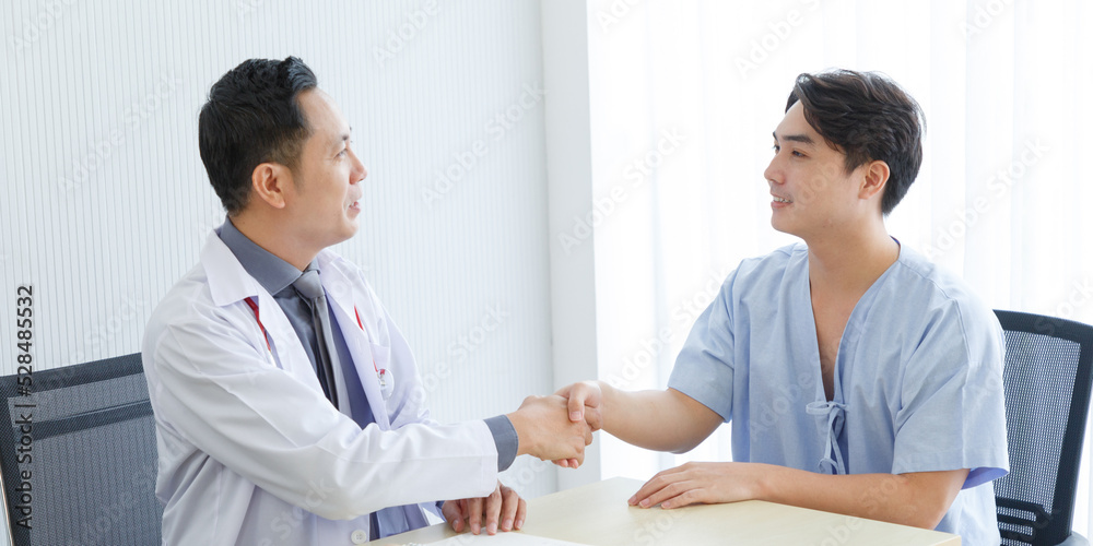 Fototapeta premium Male doctor and young male patient while consult and .explain. Doctor and patient sitting together and shake hand at table in clinic
