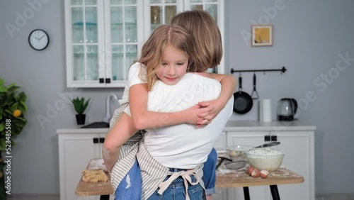 portrait of charming female child in arms of mother kisses parent on cheek smiling and looking at camera on background of kitchen at home