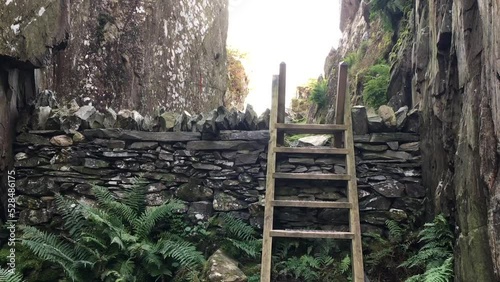 A wooden ladder stile over a dry stone wall, leading into a narrow rock ravine with mountains in the background. Llyn Idwal, Snowdonia National Park, Wales.