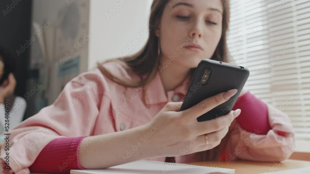 Low angle selective focus of female Caucasian high school student sitting at desk in class in morning, using smartphone Low angle selective focus of female Caucasian high school student sitting at desk in class in morning, using smartphone