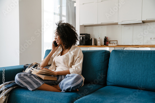 Black woman talking on cellphone and reading book at home © Drobot Dean