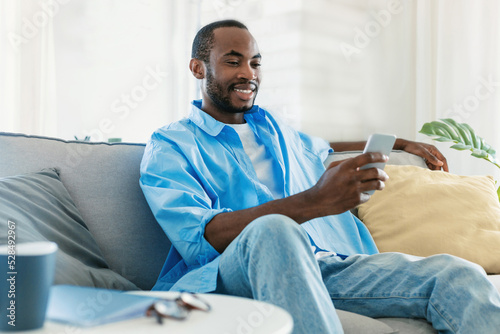 Positive black man using smartphone, scrolling news feed in social media app or texting online, sitting on sofa
