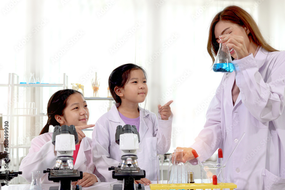 Students and teacher in lab coat having fun together while learning science experiment in laboratory. Young pretty Asian scientist showing chemical flasks to two adorable schoolgirls and girls pointin Students and teacher in lab coat having fun together while learning science experiment in laboratory. Young pretty Asian scientist showing chemical flasks to two adorable schoolgirls and girls pointin