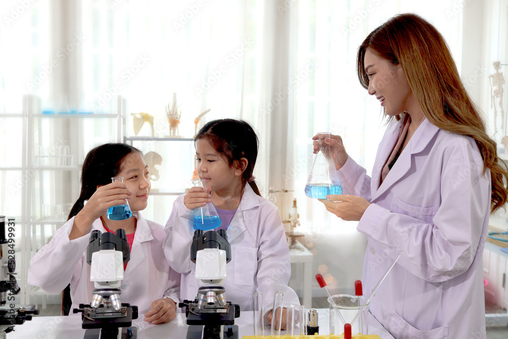 Students and teacher in lab coat have fun together while learn science experiment in laboratory. Young pretty Asian scientist showing chemical flasks to two adorable schoolgirls who look at each other Students and teacher in lab coat have fun together while learn science experiment in laboratory. Young pretty Asian scientist showing chemical flasks to two adorable schoolgirls who look at each other