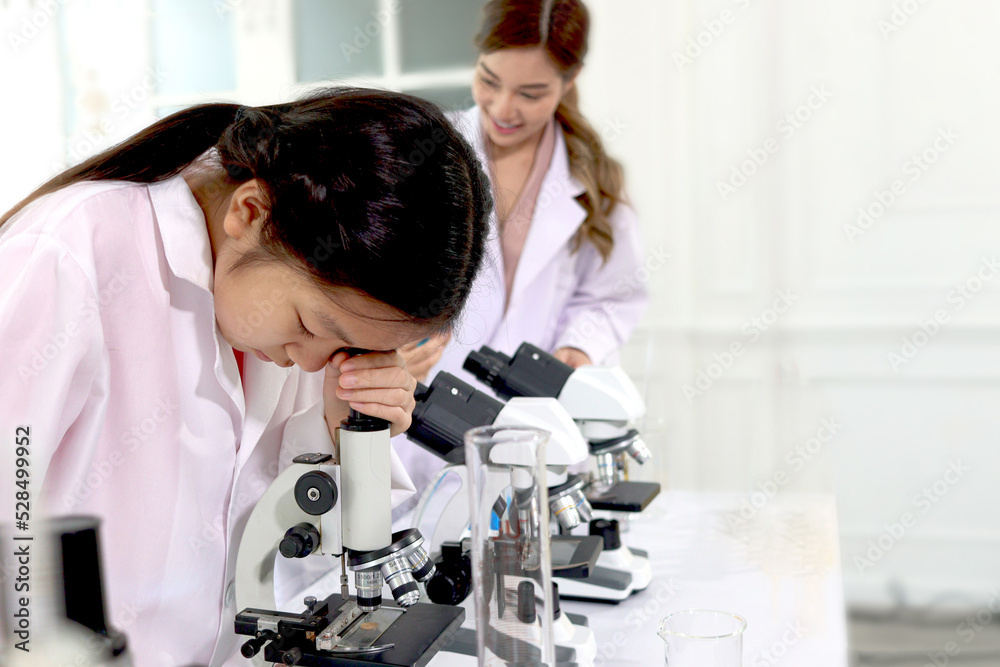 Students and teacher in lab coat have fun together, two little children students look through microscope and learn science experiment in laboratory with female teacher in classroom. Young pretty Asian Students and teacher in lab coat have fun together, two little children students look through microscope and learn science experiment in laboratory with female teacher in classroom. Young pretty Asian