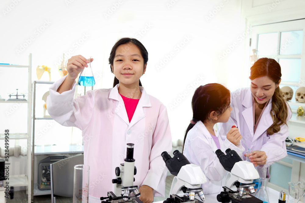 Students and teacher in lab coat have fun together while learn science experiment in laboratory. Young adorable Asian scientist kid showing blue flask with teacher and classmate as blur background. Students and teacher in lab coat have fun together while learn science experiment in laboratory. Young adorable Asian scientist kid showing blue flask with teacher and classmate as blur background.
