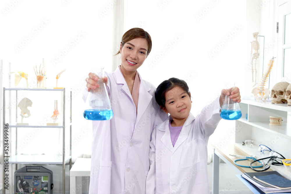 Portrait of beautiful Asian science teacher and adorable schoolgirl in lab coat standing and showing blue chemical flasks to camera in laboratory. Smiling young scientist with kid have fun together Portrait of beautiful Asian science teacher and adorable schoolgirl in lab coat standing and showing blue chemical flasks to camera in laboratory. Smiling young scientist with kid have fun together