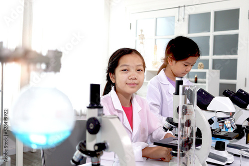 Two adorable pretty schoolgirls in lab coat doing simple science experiments, young Asian kid scientist having fun in chemistry laboratory class, little children students studying research and doing c