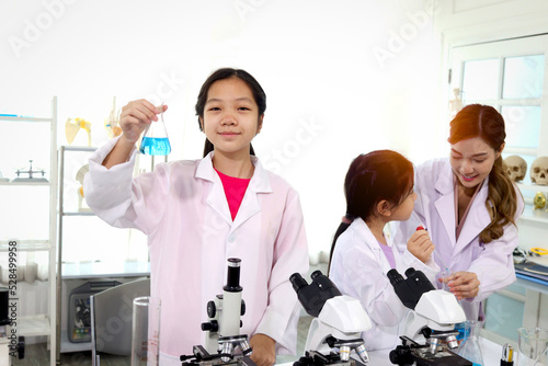 Students and teacher in lab coat have fun together while learn science experiment in laboratory. Young adorable Asian scientist kid showing blue flask with teacher and classmate as blur background.