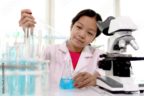 Adorable schoolgirl in lab coat doing science experiments, young scientist adding some chemical to blue flask with pipette and learning science experiment in laboratory. Asian kid using microscope, ap