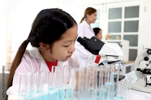 Adorable schoolgirl in lab coat doing science experiments  young scientist looking through microscope and learn science experiment in laboratory. Asian kid using apparatus and lab equipment for resear