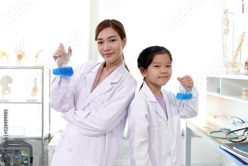 Portrait of beautiful Asian science teacher and adorable schoolgirl in lab coat holding blue chemical flasks and standing back to back with arms crossed in laboratory. Smiling young scientist with kid
