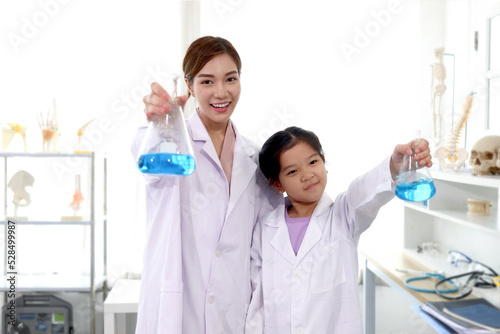 Portrait of beautiful Asian science teacher and adorable schoolgirl in lab coat standing and showing blue chemical flasks to camera in laboratory. Smiling young scientist with kid have fun together an