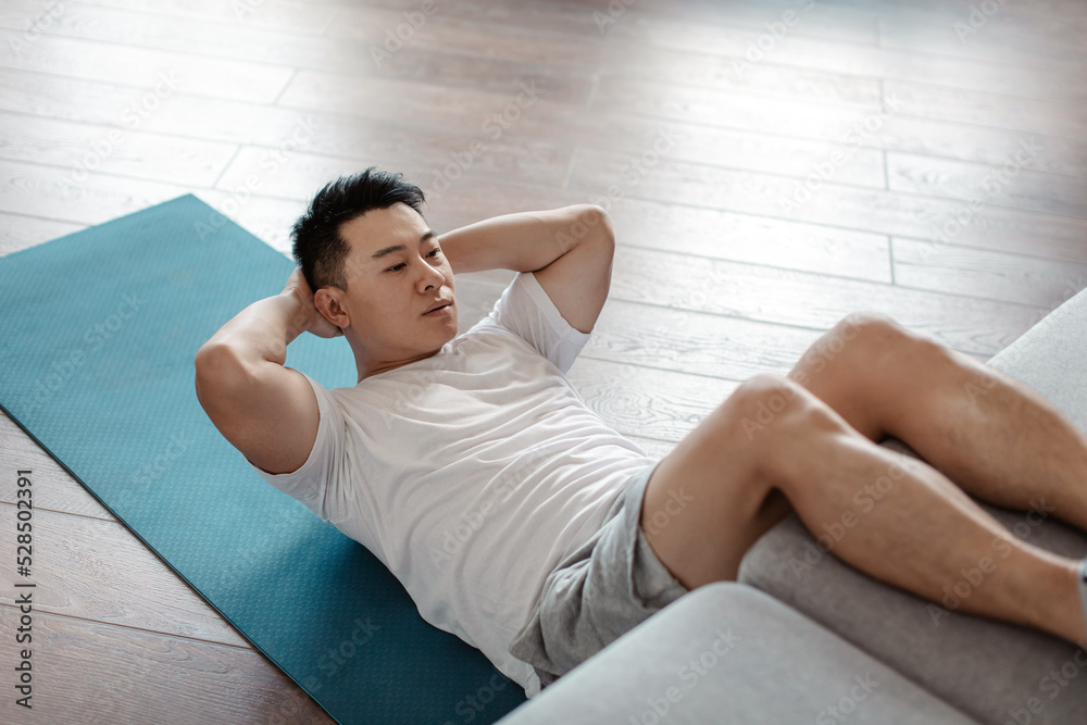 Athletic korean mature man exercising abs muscles on yoga mat, putting legs on sofa, working out at home in the morning Athletic korean mature man exercising abs muscles on yoga mat, putting legs on sofa, working out at home in the morning