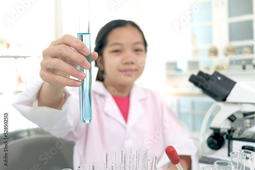 Hand of schoolgirl in lab coat showing blue chemical showing test tube to camera, kid doing science experiments, young scientist student learning science experiment in laboratory. Asian kid using micr photo