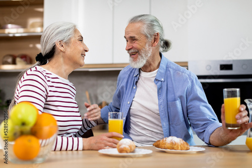 Domestic Morning. Happy Senior Spouses Chatting And Laughing During Breakfast In Kitchen