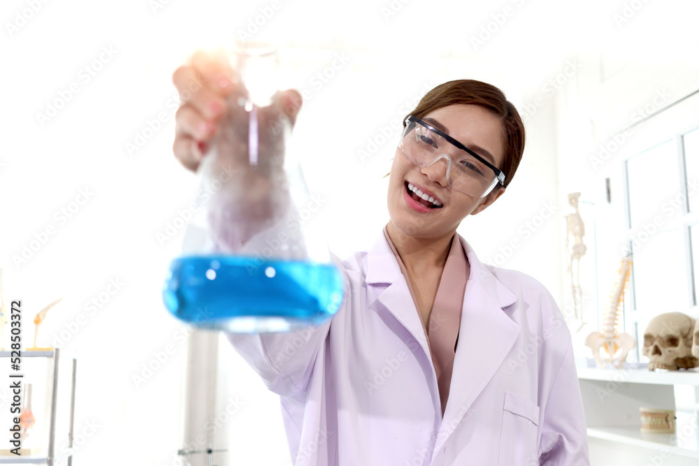 Portrait of beautiful Asian female scientist wear lab coats and safety glasses, happy smiling researcher woman standing and showing blue chemical flasks to camera in laboratory, woman working in scien Portrait of beautiful Asian female scientist wear lab coats and safety glasses, happy smiling researcher woman standing and showing blue chemical flasks to camera in laboratory, woman working in scien