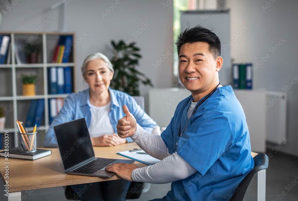 Cheerful adult asian male doctor showing thumb up sign with hand and consultation elderly female patient
