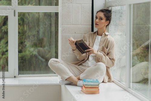 Young beautiful girl sitting on windowsill, holding and reading book