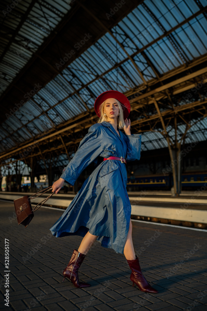 Fashionable confident blonde woman wearing dark red hat, blue trench coat, belt, leather ankle boots, holding stylish bag. Female fashion conception. Outdoor full-length portrait