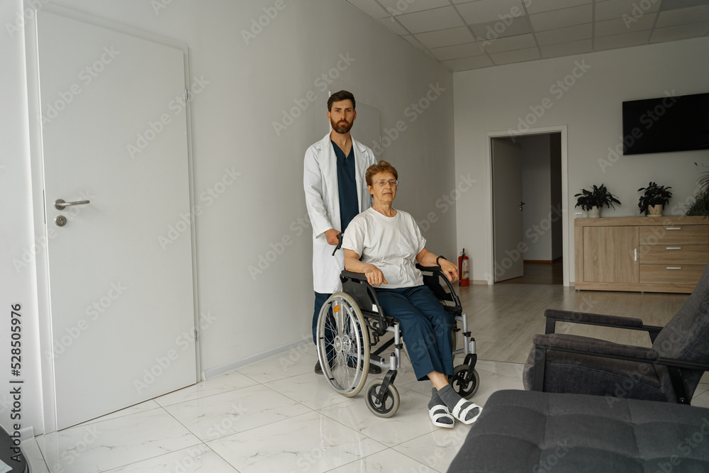 Professional male doctor carrying patient on wheelchair in medicine clinic hall. High quality photo Professional male doctor carrying patient on wheelchair in medicine clinic hall. High quality photo